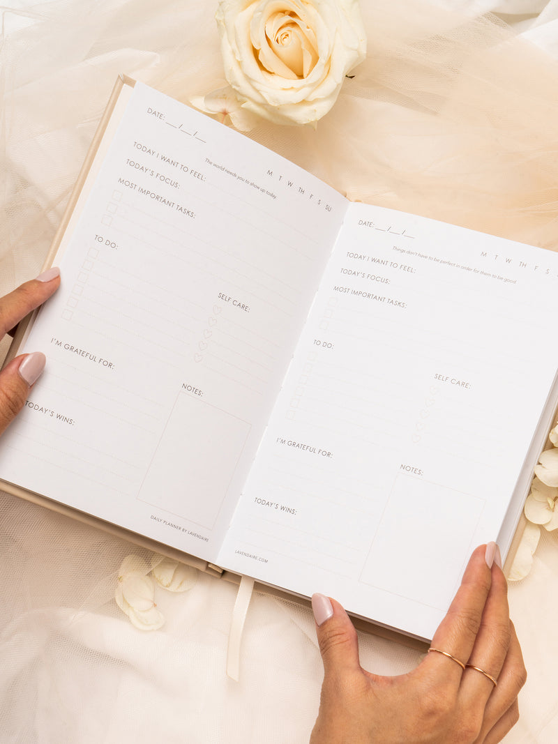 Hands holding Lavendaire Daily Planner open on beige tulle surrounded by flowers