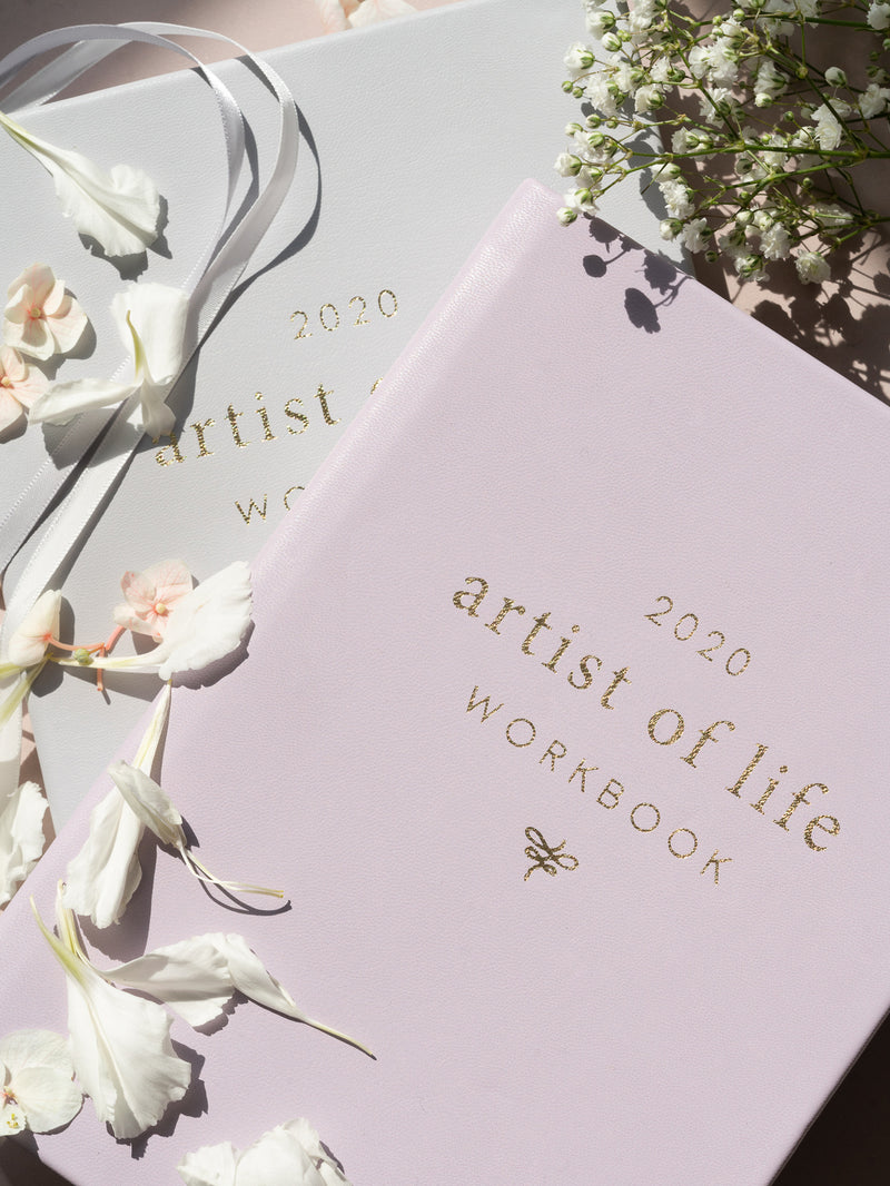 lavender artist of life workbook on top of grey workbook with white flowers, petals, and ribbons resting on top