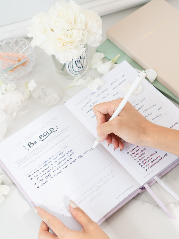 Person writing in lavender artist of life workbook next to Pastel Notebook, Daily Planner, jar with white flowers