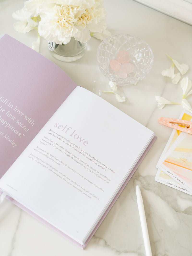 lavender artist of life workbook open to self love section, next to jar of white flowers, bowl with pink crystals, flower petals, tarot cards, pink hair clip, and white pen