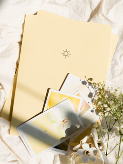 two yellow pastel notebooks on an off-white fabric background, tarot cards resting on top, next to white flowers and perfume bottle