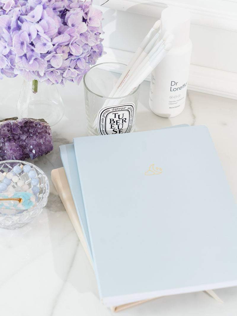 two blue pastel notebooks stacked on top of daily planner on marble surface, next to bowl containing beaded bracelets and blue hair clip, amethyst crystal, jar of lavender flowers, jar of white paint brushes