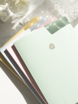 green, pink, blue, and yellow notebooks stacked on top of each other, fanned out, next to crystal container and jar of white flowers